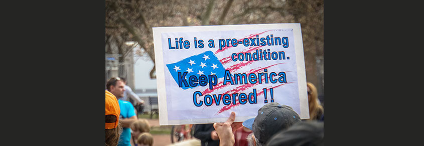 A photo of a sign from a protest that says, "Life is a pre-existing condition. Keep America Covered!!"