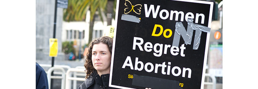 A photo of a woman holding a sign that says "Women Don't Regret Abortion"