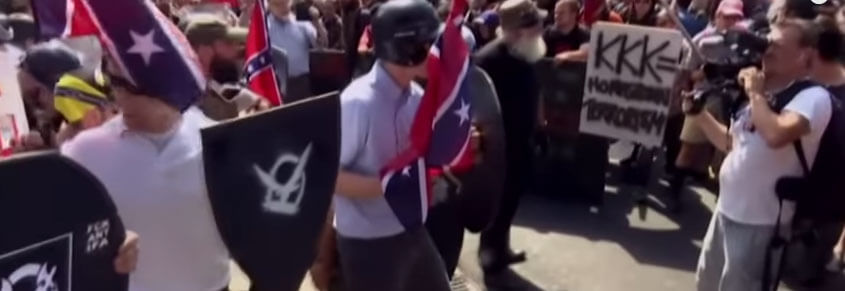 A photo from the Unite the Right rally. White men are holding signs with white supremacist symbols and Confederate flags.