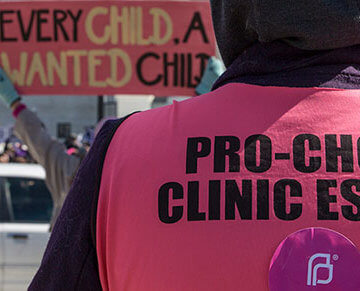 A picture of someone wearing a shirt that says "Pro-Choice Clinic Escort," with anti-abortion protestors around them.