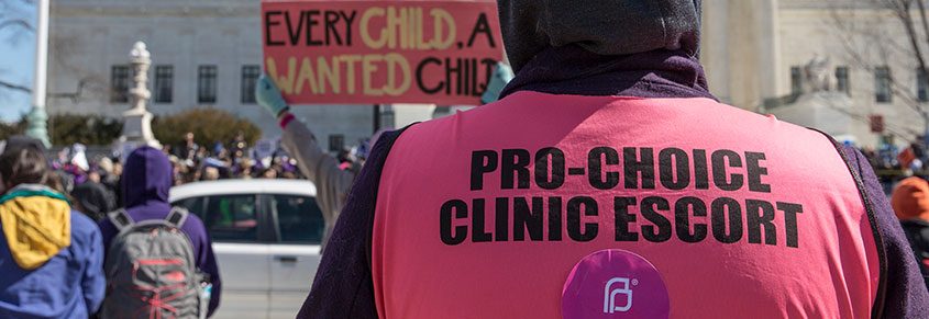 A picture of someone wearing a shirt that says "Pro-Choice Clinic Escort," with anti-abortion protestors around them.