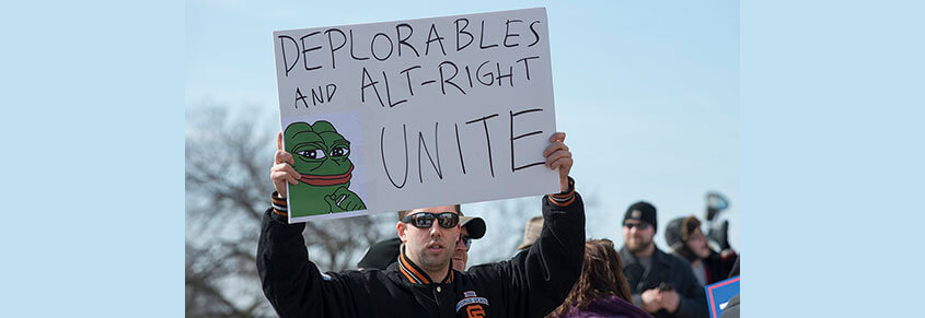 A photo of a white man holding a sign that says "Deplorables and Alt-Right Unite" with a photo of Pepe the Frog on it.