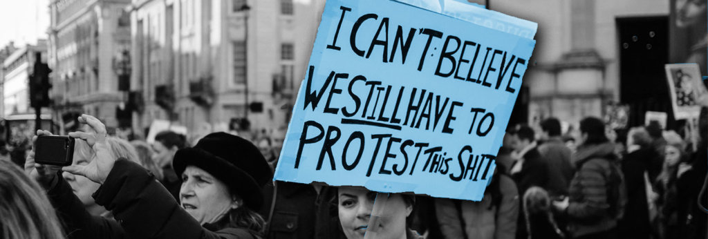 A photo from a protest. One woman is carrying a sign that says, "I can't believe we still have to protest this shit."