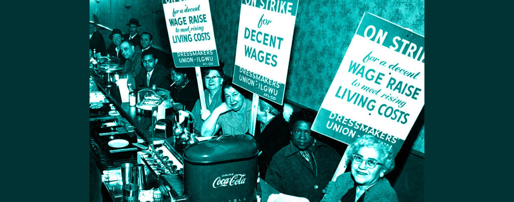 A photo from the earl 20th century of women holding pro-union signs, including one that says "On Strike for a decent Wage Raise to need rising Living Costs"