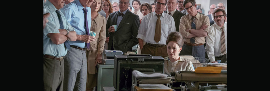 A still from the movie "The Post." Mostly male journalists surround a female journalist who is working at her desk.