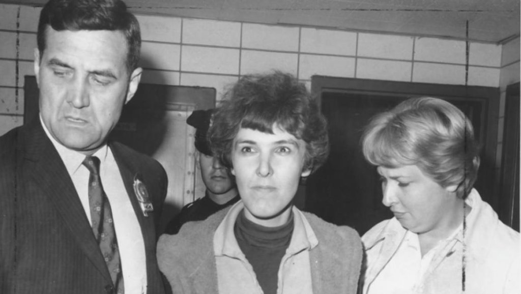 A photo of Valerie Solanas after she had been arrested. A police officer is in the background.
