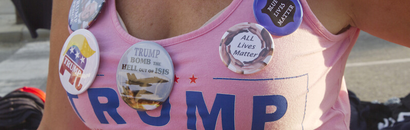 A white woman wearing a Trump tank top with buttons like, "Blue Lives Matter," "All Lives Matter," and "Trump Bomb the Hell Out of Isis"