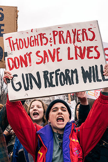 A photo from a protest of teens fighting for gun control. Two visible signs say "Fear has no place in school" and "Thoughts and prayers don't save lives. Gun reform will."