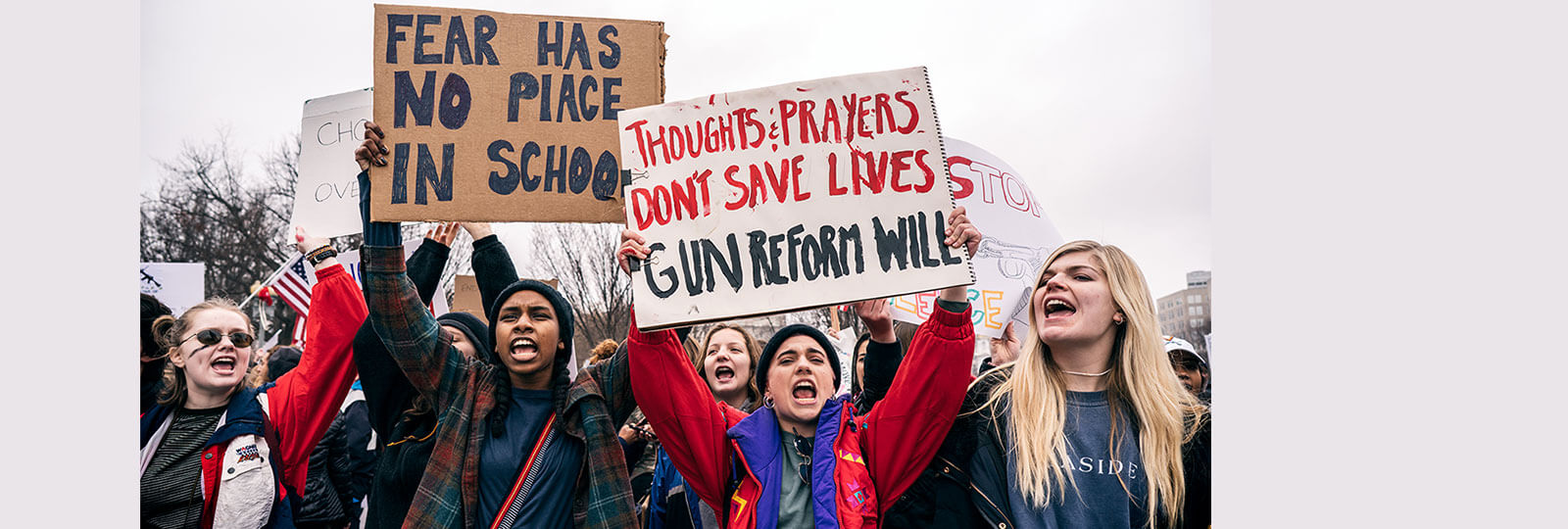 A photo from a protest of teens fighting for gun control. Two visible signs say "Fear has no place in school" and "Thoughts and prayers don't save lives. Gun reform will."