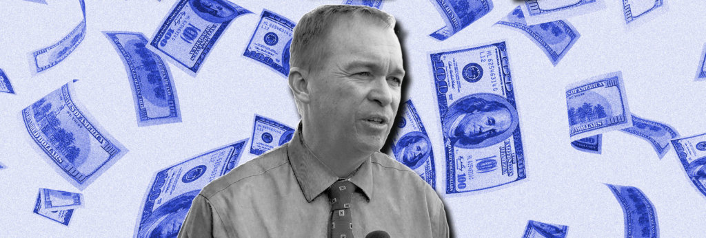 A photo of Mick Mulvaney will a 100 dollar bills flying behind him.