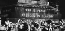 A photo of people in front of a monument that says "War is peace. Freedom is slavery."