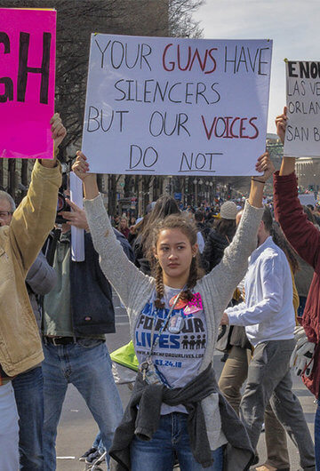 People protesting against gun violence. Signs have messages like "Enough" and "Your Guns Have Silencers But Our Voices Do Not"