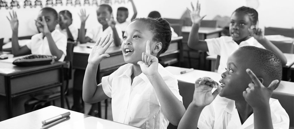 A photo of Black children raising their hands in a classroom.