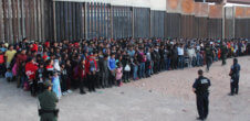 A photo of at least 100 youths lined up against fences with a few armed CBP officers.