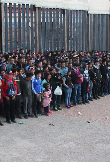 A photo of at least 100 youths lined up against fences with a few armed CBP officers.
