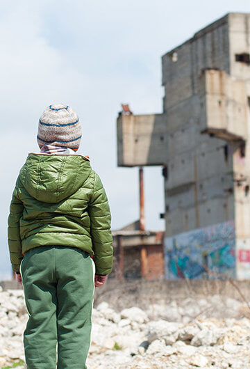 A photo of a young refugee child in a winter jacket in front of some abandoned structures.
