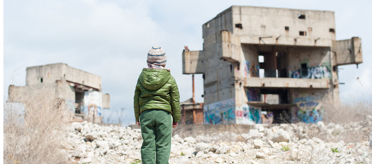 A photo of a young refugee child in a winter jacket in front of some abandoned structures.