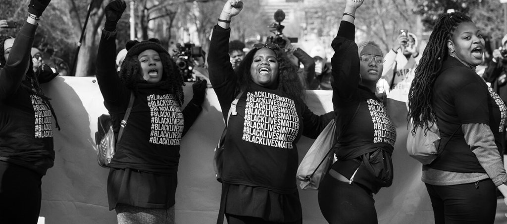 A photo of a Black Lives Matter protest. Five women in front are wearing #BlackLivesMatter shirts, and four out of five are raising their fists.