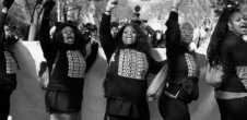 A photo of a Black Lives Matter protest. Five women in front are wearing #BlackLivesMatter shirts, and four out of five are raising their fists.