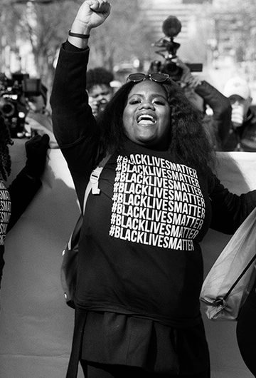 A photo of a Black Lives Matter protest. Five women in front are wearing #BlackLivesMatter shirts, and four out of five are raising their fists.