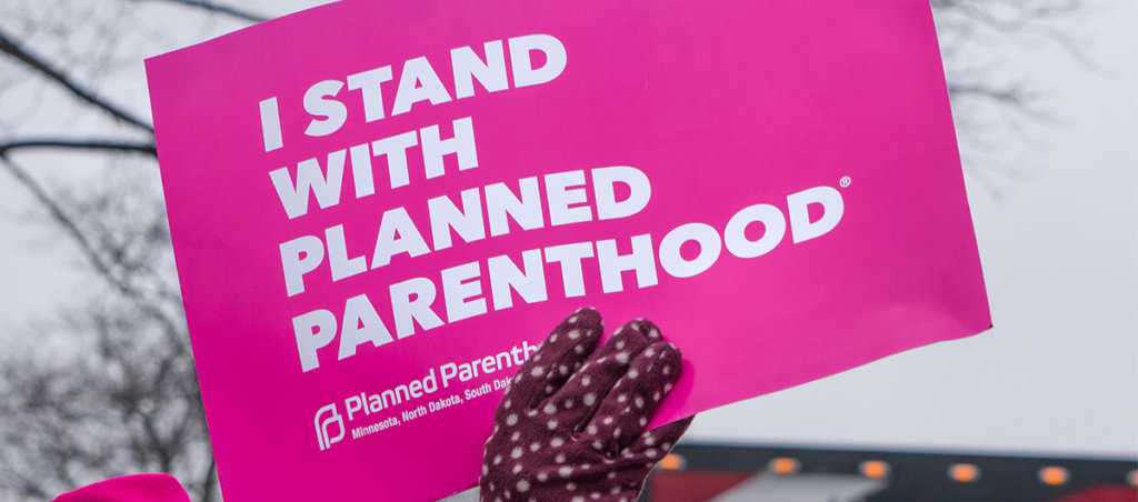 A hand in a mitten holding a sign that says, "I Stand With Planned Parenthood"