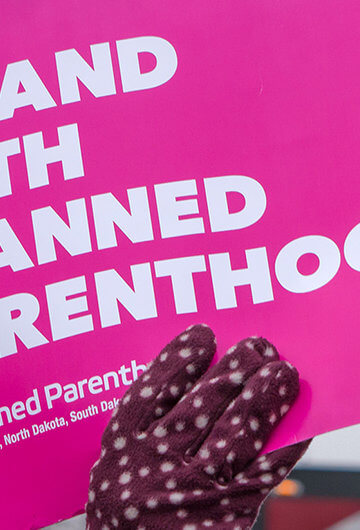 A hand in a mitten holding a sign that says, "I Stand With Planned Parenthood"