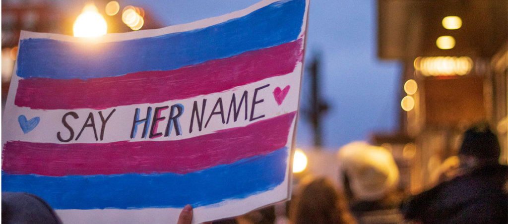 Photo from a protest that says "Say Her Name" with blue and pink on it, which are colors of the trans visibility flag.