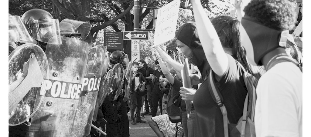 A black and white photos of police in riot gear and protestors holding up signs,