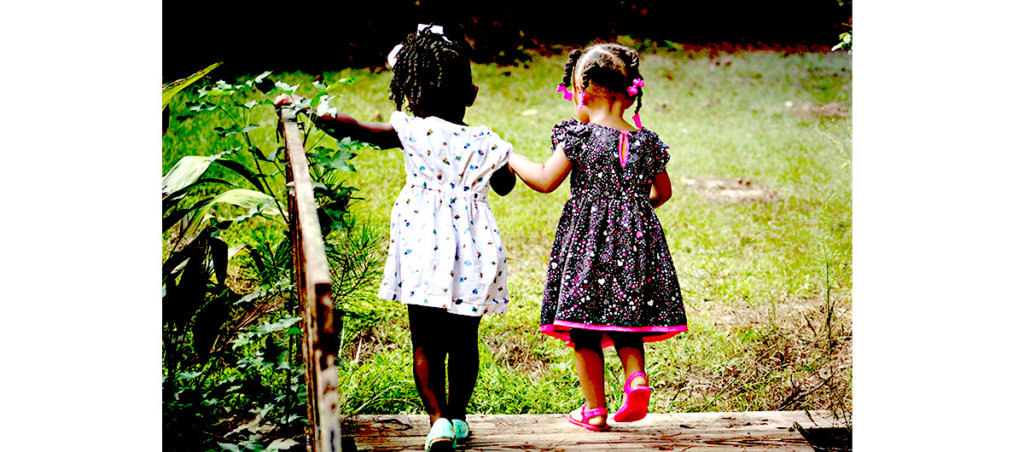 An image of two Black girls in dresses holding hands and walking in the other direction in a grassy area.