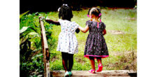 An image of two Black girls in dresses holding hands and walking in the other direction in a grassy area.