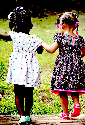 An image of two Black girls in dresses holding hands and walking in the other direction in a grassy area.