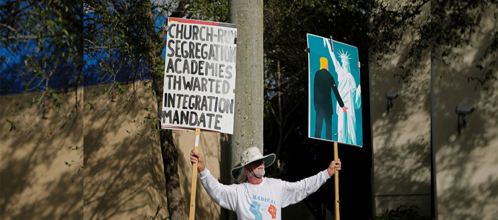 A photo of a man holding two signs. First sign says "Church-run segregation academies thwarted. Integration Mandate." Second sign in an illustration of Trump groping the Statue of Liberty.