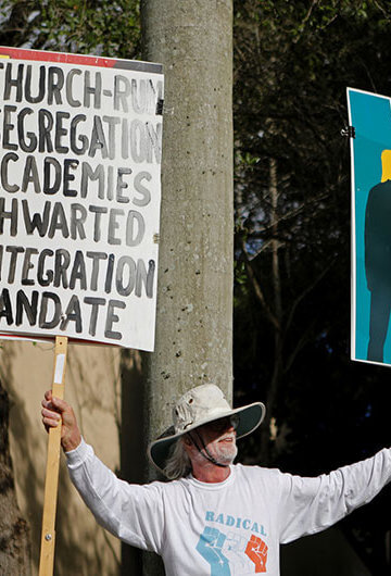 A photo of a man holding two signs. First sign says "Church-run segregation academies thwarted. Integration Mandate." Second sign in an illustration of Trump groping the Statue of Liberty.