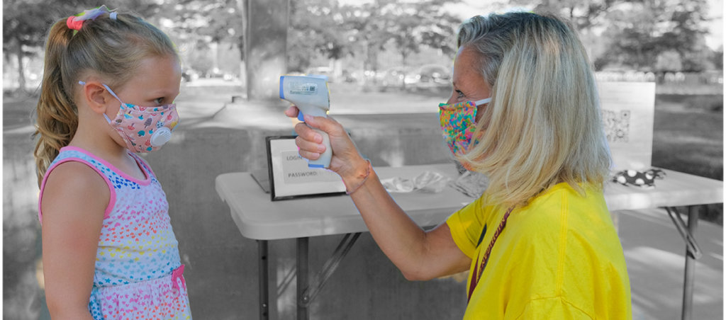 A photo of a white woman with blonde hair taking the temperature of a kid with blonde hair. Both are wearing masks.