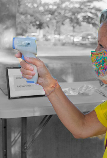 A photo of a white woman with blonde hair taking the temperature of a kid with blonde hair. Both are wearing masks.