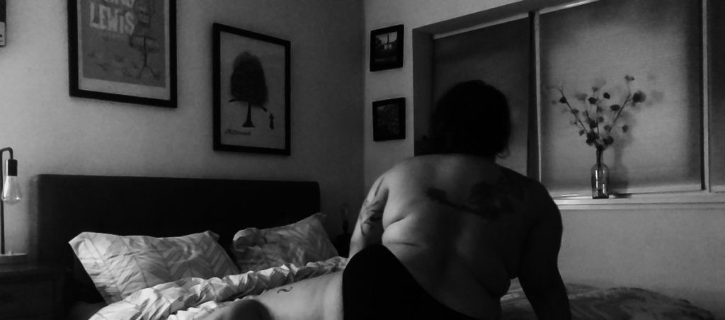 A black and white photo of someone on the bed topless facing the other direction.