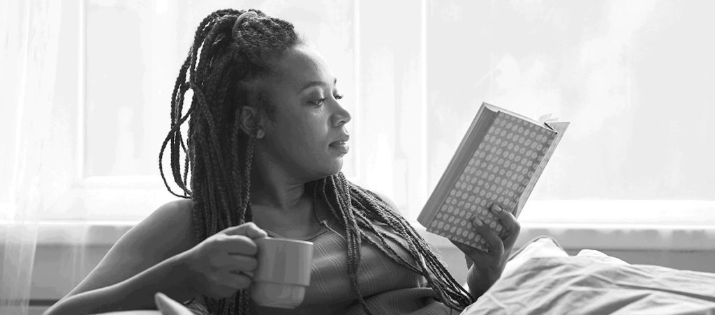 Black woman holding a coffee mug and reading a book while in a reclined position