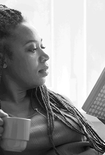 Black woman holding a coffee mug and reading a book while in a reclined position