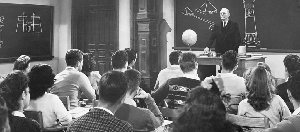 A white male professor speaking at the front of a university classroom. Students are sitting in desks facing the processor.