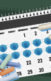 a calendar with covid masks on the dates. On the lower left corner is a pile of chalk. On the upper right corner, a pile of pills