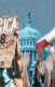 People's hands are visible at a protest, with a megaphone and a sign that says "America 2020 Ain't Shit Changed" with the number 1968 crossed out. The White House is in the background.