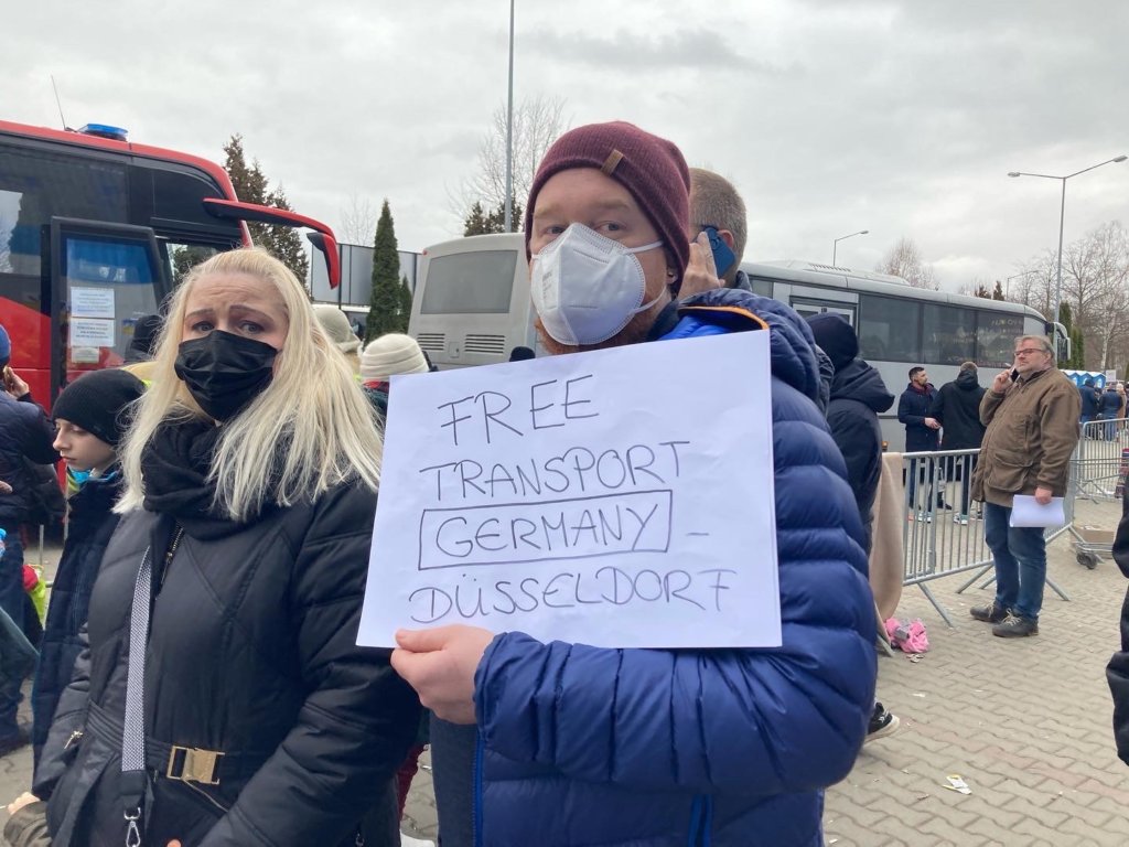 A white man in a blue jacket and red hat holding a sign that says "Free Transport Germany" outside – he is surrounded by people and a few buses