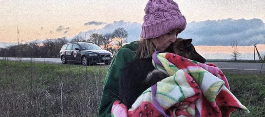 A white woman with blonde hair in a green jacket and pink hat holding a dog wrapped in a blanket. She is outside standing on some grass.