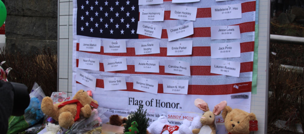 An American flag with names on it honoring people who died at the Sandy Hook shooting. There are also stuffed animals near the flag.