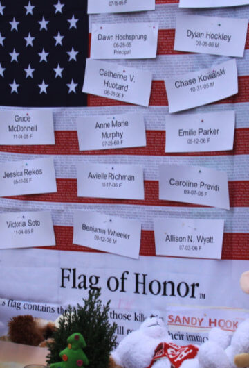 An American flag with names on it honoring people who died at the Sandy Hook shooting. There are also stuffed animals near the flag.