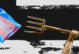 A man's arm holding a pitchfork in the direction of a trans flag.